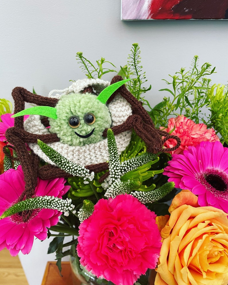 K&ouml;nnte beinhalten: Eine gr&uuml;ne geh&auml;kelte Spinne mit Glitz Augen und einem l&auml;chelnden Mund sitzt in einer Vase mit rosa, orangefarbenen und wei&szlig;en Blumen.