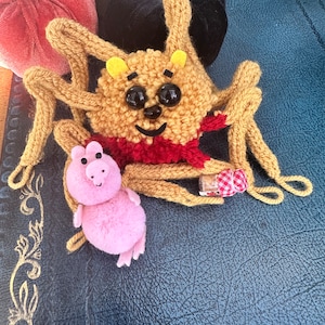 May include: A brown knitted spider with large black eyes and a red and white scarf. The spider is holding a small pink pig plush toy.
