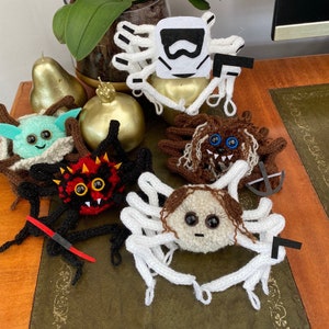 K&ouml;nnte beinhalten: Eine Sammlung von sieben geh&auml;kelten Amigurumi-Figuren, darunter ein wei&szlig;er Stormtrooper, ein brauner Chewbacca, ein gr&uuml;ner Yoda, eine rote Spinne, eine wei&szlig;e Spinne, eine braune Spinne und eine wei&szlig;e Spinne mit einem braunen Gesicht. Die Figuren sitzen alle auf einem Holztisch.
