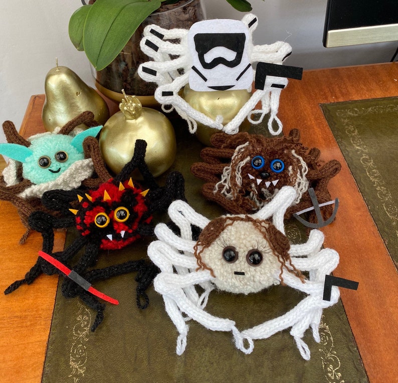 K&ouml;nnte beinhalten: Eine Sammlung von sieben geh&auml;kelten Amigurumi-Figuren, darunter ein wei&szlig;er Stormtrooper, ein brauner Chewbacca, ein gr&uuml;ner Yoda, eine rote Spinne, eine wei&szlig;e Spinne, eine braune Spinne und eine wei&szlig;e Spinne mit einem braunen Gesicht. Die Figuren sitzen alle auf einem Holztisch.