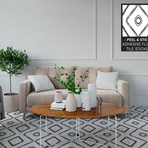 Op de afbeelding: Een woonkamer met een beige bank, twee planten en een houten salontafel. De vloer is bedekt met zwart-witte peel-and-stick vloertegelstickers. De tekst op de ingelijste poster luidt "Peel & Stick - Adhesive Floor Tile Sticker."