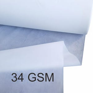 Peut inclure: Gros plan sur un rouleau de tissu blanc avec une couche bleu-gris visible en dessous. Le texte "34 GSM" est imprimé en noir sur la couche inférieure du tissu. Le tissu semble doux et léger.