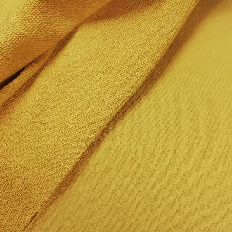 K&ouml;nnte beinhalten: Nahaufnahme eines gelben Stoffes mit strukturierter Oberfl&auml;che. Der Stoff ist in der Mitte gefaltet und bildet eine diagonale Linie.