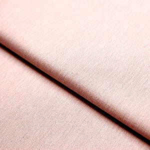 Pode incluir: Close-up de uma seção de tecido rosa. O material tem uma trama fina e texturizada, e a cor é um rosa suave e discreto. O tecido está dobrado, criando sombras e destacando a textura.