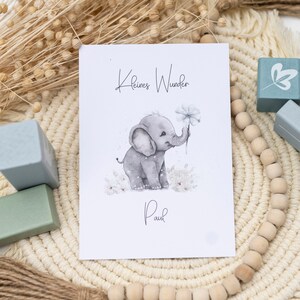 Pode incluir: Um cart&atilde;o branco com o texto "Kleines Wunder" e "Paul" em escrita preta. Uma ilustra&ccedil;&atilde;o em aquarela de um elefante cinza segurando uma flor branca est&aacute; no centro do cart&atilde;o. O cart&atilde;o est&aacute; sobre um tapete tecido bege com contas de madeira e blocos de madeira azuis.