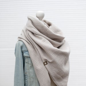 Könnte beinhalten: Ein hellgrauer Baumwollschal, der über eine Schaufensterpuppe mit Jeansjacke gelegt ist. Der Schal ist aus einem weichen, strukturierten Stoff und hat ein kleines braunes Etikett mit einem Stern.