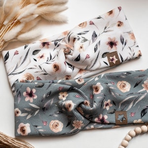 Könnte beinhalten: Zwei Blumenmuster-Stirnbänder, eines weiß mit braunen und rosa Blumen und eines grau mit braunen und rosa Blumen. Beide Stirnbänder haben ein braunes Leder-Etikett mit einem Stern.