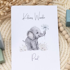 Pode incluir: Um cart&atilde;o branco com o texto "Kleines Wunder" e o nome "Paul" escrito em it&aacute;lico. Uma ilustra&ccedil;&atilde;o em aquarela de um elefante cinza segurando uma flor branca est&aacute; no centro do cart&atilde;o.