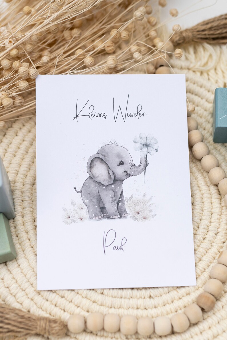 Pode incluir: Um cart&atilde;o branco com o texto "Kleines Wunder" e o nome "Paul" escrito em it&aacute;lico. Uma ilustra&ccedil;&atilde;o em aquarela de um elefante cinza segurando uma flor branca est&aacute; no centro do cart&atilde;o.