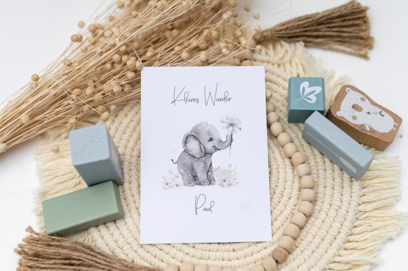 Pode incluir: Um cart&atilde;o branco com o texto "Kleines Wunder" e "Paul" em letras pretas. Uma ilustra&ccedil;&atilde;o em aquarela de um elefante cinza segurando uma flor est&aacute; no centro do cart&atilde;o. O cart&atilde;o &eacute; cercado por blocos de madeira e um tapete tecido bege.