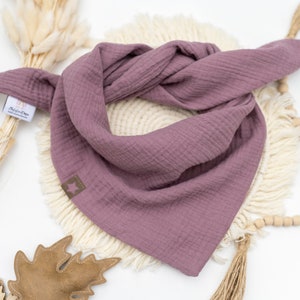 May include: A mauve-colored cotton bandana with frayed edges. The bandana is folded and displayed on a white surface with a wooden bead garland and a brown leaf.