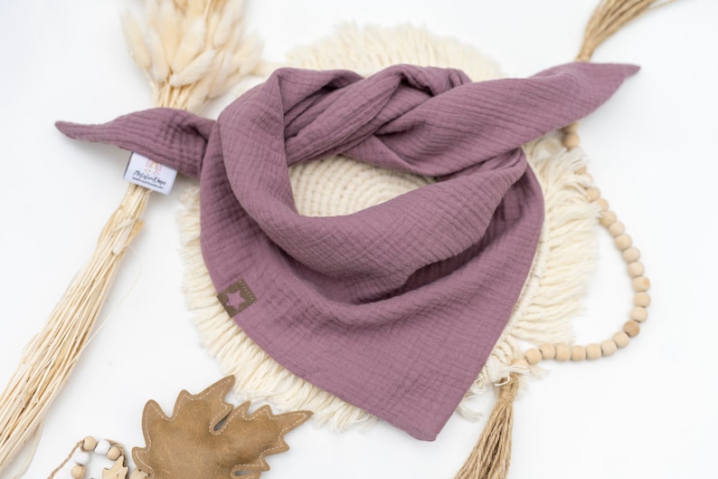 May include: A mauve-colored cotton bandana with frayed edges. The bandana is folded and displayed on a white surface with a wooden bead garland and a brown leaf.