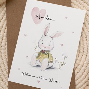 Op de afbeelding: Wenskaart met een handgetekende illustratie van een witte konijn in een groene jas, met de naam "Amelia" boven een roze hart. De tekst "Willkommen kleines Wunder" staat eronder. De kaart ligt op een bruine envelop.