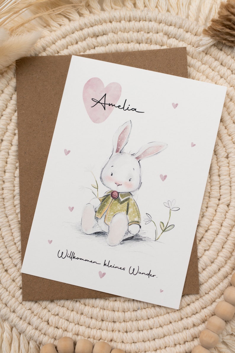 Op de afbeelding: Wenskaart met een handgetekende illustratie van een witte konijn in een groene jas, met de naam "Amelia" boven een roze hart. De tekst "Willkommen kleines Wunder" staat eronder. De kaart ligt op een bruine envelop.