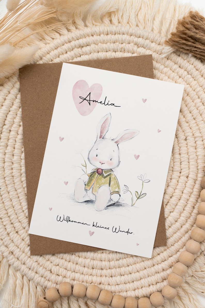 Op de afbeelding: Een wenskaart met een aquarel illustratie van een witte konijn met een groene jas. De kaart bevat de naam "Amelia" boven een roze hart en de zin "Willkommen kleines Wunder."
