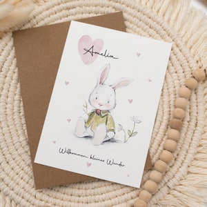 Op de afbeelding: Wenskaart met een aquarel illustratie van een konijn met een groene jas, de naam "Amelia" en de tekst "Willkommen kleines Wunder." De kaart ligt op een bruine envelop, met een macram&eacute; placemat op de achtergrond.