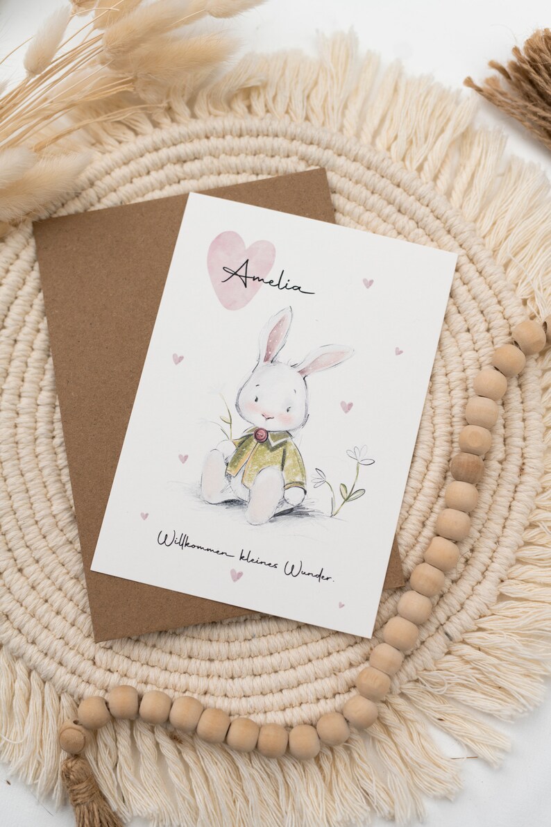 Op de afbeelding: Wenskaart met een aquarel illustratie van een konijn met een groene jas, de naam "Amelia" en de tekst "Willkommen kleines Wunder." De kaart ligt op een bruine envelop, met een macram&eacute; placemat op de achtergrond.