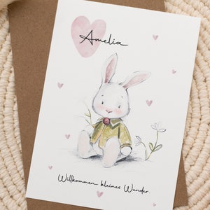 Op de afbeelding: Een wenskaart met een aquarel illustratie van een witte konijn in een groene jas, met de naam "Amelia" erboven. De kaart bevat de tekst "Willkommen kleines Wunder" en kleine roze hartjes.