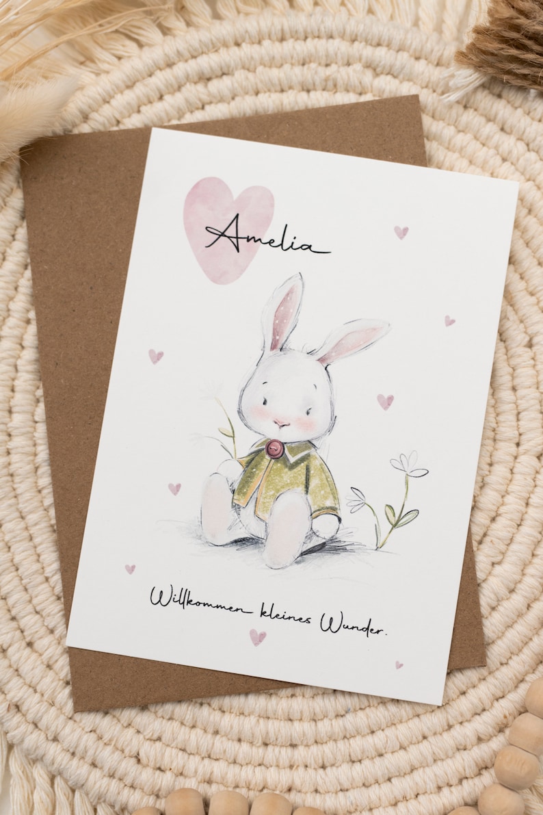 Op de afbeelding: Een wenskaart met een aquarel illustratie van een witte konijn in een groene jas, met de naam "Amelia" erboven. De kaart bevat de tekst "Willkommen kleines Wunder" en kleine roze hartjes.