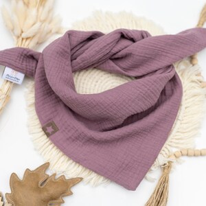 May include: A mauve-colored cotton bandana with frayed edges. The bandana is folded and displayed on a white surface with a wooden bead garland and a brown leaf.
