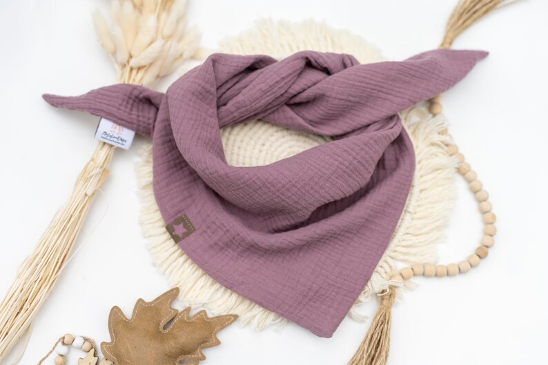 May include: A mauve-colored cotton bandana with frayed edges. The bandana is folded and displayed on a white surface with a wooden bead garland and a brown leaf.