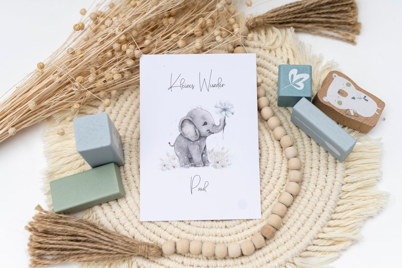 Pode incluir: Um cart&atilde;o branco com o texto "Kleines Wunder" e "Paul" em escrita preta. Uma ilustra&ccedil;&atilde;o em aquarela de um elefante cinza segurando uma flor branca est&aacute; no centro do cart&atilde;o. O cart&atilde;o est&aacute; sobre um tapete tecido bege com contas de madeira e blocos de madeira azuis.