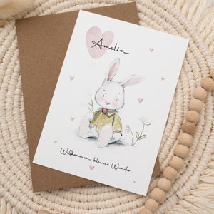 Op de afbeelding: Een wenskaart met een aquarel illustratie van een witte konijn met een groene jas, met de naam "Amelia" boven een roze hart. De kaart bevat de tekst "Willkommen kleines Wunder" en ligt op een geweven placemat.