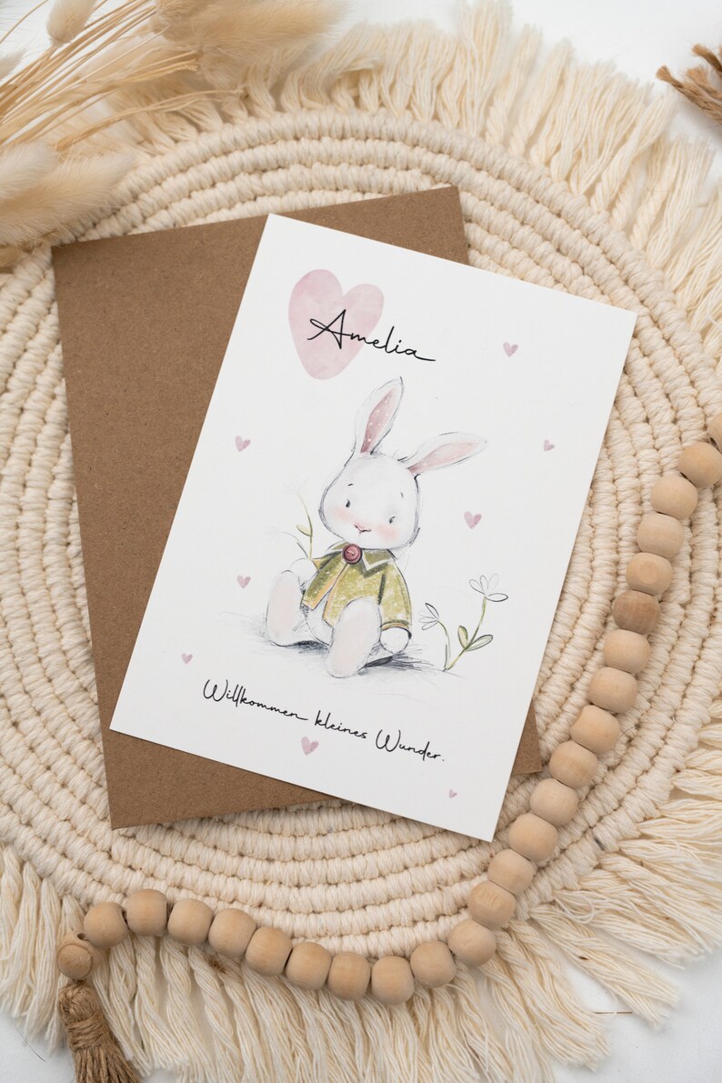 Op de afbeelding: Een wenskaart met een aquarel illustratie van een witte konijn met een groene jas, met de naam "Amelia" boven een roze hart. De kaart bevat de tekst "Willkommen kleines Wunder" en ligt op een geweven placemat.