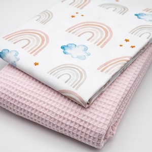 Könnte beinhalten: Eine rosa Waffel-Decke, die auf einem weißen Stoff mit einem sich wiederholenden Muster aus Aquarell-Regenbögen und Wolken gefaltet ist. Der Stoff hat ein dezentes goldenes Glitzermuster.