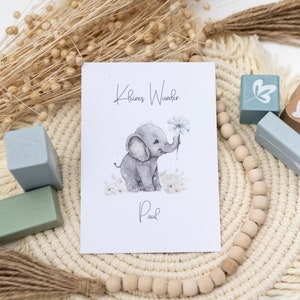 Pode incluir: Um cart&atilde;o branco com o texto "Kleines Wunder" e uma ilustra&ccedil;&atilde;o em aquarela de um elefante cinza segurando uma flor. O texto "Paul" est&aacute; escrito abaixo do elefante. O cart&atilde;o &eacute; cercado por contas de madeira e um macram&ecirc; bege.