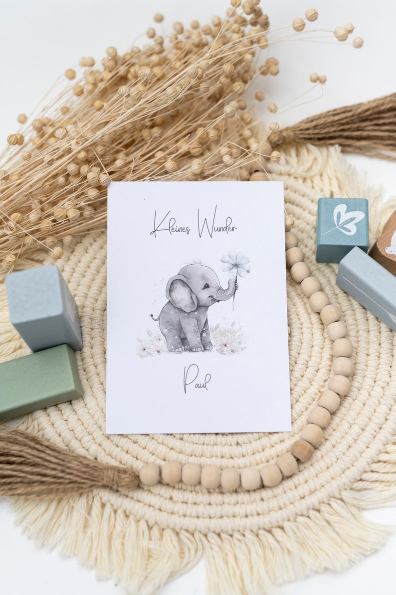 Pode incluir: Um cart&atilde;o branco com o texto "Kleines Wunder" e uma ilustra&ccedil;&atilde;o em aquarela de um elefante cinza segurando uma flor. O texto "Paul" est&aacute; escrito abaixo do elefante. O cart&atilde;o &eacute; cercado por contas de madeira e um macram&ecirc; bege.
