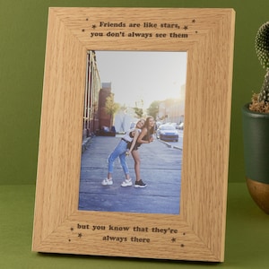 May include: A light oak photo frame with the inscription "Friends are like stars, you don't always see them, but you know that they're always there." The frame contains a photograph of two people. A cactus in a green and gold pot is to the right.