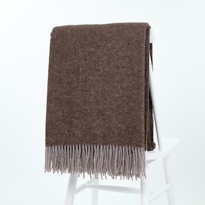 May include: A rectangular throw blanket in a solid dark brown color with a textured, woven appearance. The blanket features a fringe trim along the bottom edge in a lighter, muted grey tone, creating a distinct contrast against the brown fabric.