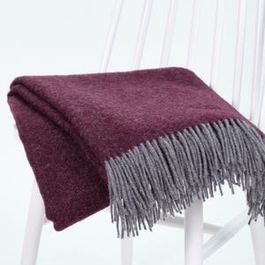 Op de afbeelding: Een gevouwen bordeauxrode deken met grijze franjes ligt over de rugleuning van een witte houten stoel. De deken, gemaakt van een zacht, getextureerd materiaal, is ideaal voor het toevoegen van warmte en stijl.