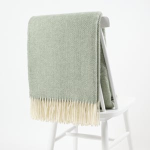 Op de afbeelding: Een zachte, groene deken met een herringbone patroon en een witte franje. De deken is over de rugleuning van een witte houten stoel gelegd.