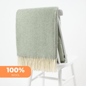 Può includere: Una coperta in lana verde salvia con motivo a spina di pesce e frange color crema è drappeggiata su una sedia bianca. L'etichetta indica "100% WOOL" in un riquadro arancione.