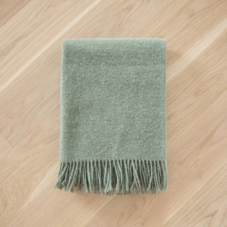 May include: A soft, sage green throw blanket with a fringe border. The blanket is folded and laid flat on a light wood floor.