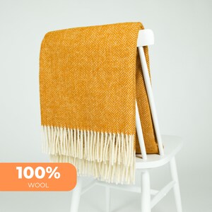 May include: Mustard yellow herringbone wool throw with cream fringe, draped over a white chair. The blanket is labelled "100% WOOL" in an orange box. The blanket is made of 100% wool.