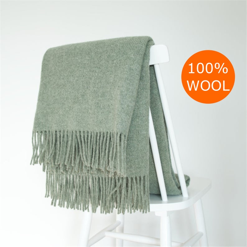 May include: A soft, sage green throw blanket with fringe on the bottom edge. The blanket is draped over a white wooden chair. The text "100% WOOL" is printed in orange on a white circle.