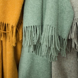 May include: Close-up of three soft, fringed throws in shades of mustard yellow, seafoam green, and grey. The throws are made of a textured material, likely wool or a similar fabric, and are folded to display their fringed edges.