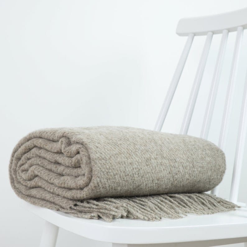 May include: A soft, beige throw blanket with fringe is folded and draped over a white chair. The blanket has a subtle herringbone pattern.