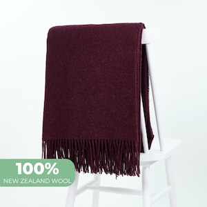 Op de afbeelding: Een bordeauxrode wollen deken met franjes op een witte stoel. De deken is gemaakt van 100% Nieuw-Zeelandse wol.