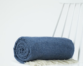 Puede incluir: Una manta azul enrollada con un sutil patrón de espiga y flecos de color crema en los extremos. La manta descansa sobre una silla de madera blanca contra un fondo blanco liso, mostrando su tejido texturizado y su color azul intenso.