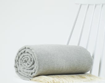 May include: A light gray, tightly rolled throw blanket featuring a soft, solid-colored texture and a fringe edge with cream-colored tassels. The blanket is positioned horizontally, showcasing its cylindrical shape and smooth fabric surface.