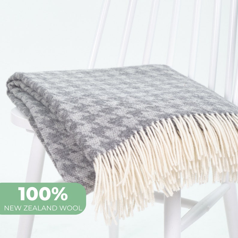 Może przedstawiać: Szara i biała wełniana narzuta w pepitkę z kremowymi frędzlami jest narzucona na białym krześle. Na narzucie widnieje napis "100% New Zealand Wool".
