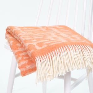 May include: A folded throw blanket featuring a vibrant orange and white abstract pattern. The blanket is finished with long, cream-colored fringe along the edge, providing a textured appearance to the woven fabric design throughout the piece.