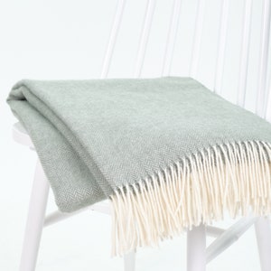 Könnte beinhalten: Eine hellgrüne Decke mit Fischgrätenmuster und cremefarbenen Fransen liegt über einem weißen Stuhl. Die Decke ist gefaltet und wirkt weich und gemütlich. Der Hintergrund ist hellweiß.