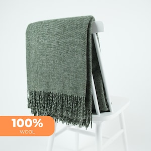 Può includere: Una coperta in lana verde intrecciata con frange, drappeggiata sullo schienale di una sedia bianca. La coperta è di un colore verde tenue e ha un aspetto strutturato. Le parole "100% WOOL" sono in un riquadro arancione.
