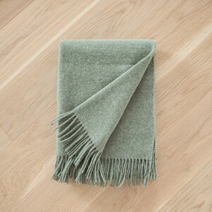 May include: A soft, sage green throw blanket with fringe on all sides. The blanket is folded in half and laid on a wood floor.