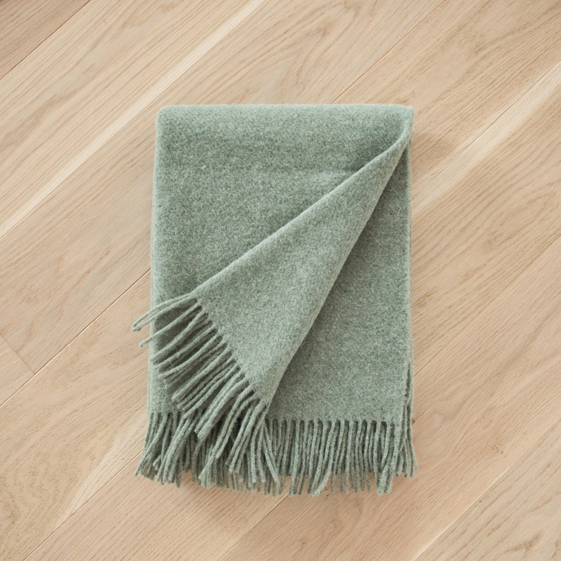 May include: A soft, sage green throw blanket with fringe on all sides. The blanket is folded in half and laid on a wood floor.